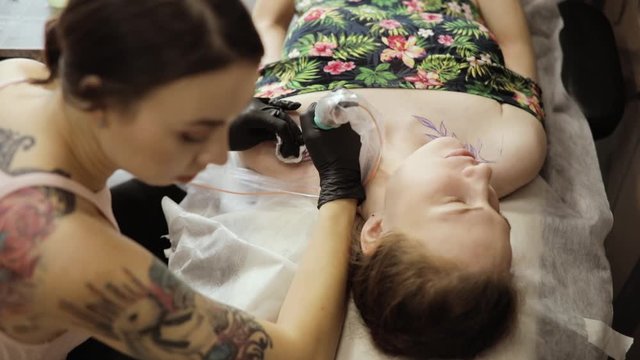 Creating body image in tattoo salon. Tattoo artist making tattoo picture with leaves at studio for young woman, top view. She uses tattooing machine making her job. Wiping skin with cotton pad.