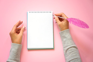Obraz premium Woman's hands holding pen with feather and notepad as mockup for your design. Pink background, flat lay style.