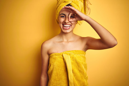 Young beautiful woman wearing a shower towel after bath over yellow isolated background doing ok gesture with hand smiling, eye looking through fingers with happy face.