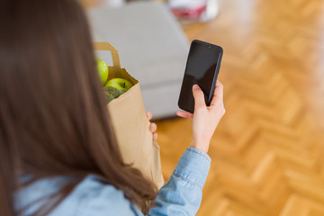 Young woman holding a paper bag full of fresh groceries and using smartphone app for supermarket delivery
