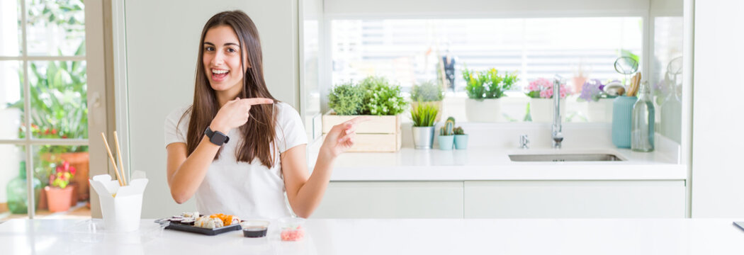 Wide angle picture of beautiful young woman eating asian sushi from delivery smiling and looking at the camera pointing with two hands and fingers to the side.