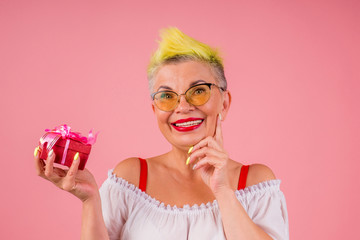 caucasian woman wear sunhlasses with stylish yellow dyed hair holding gift bow box in pink studio background