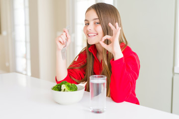 Beautiful young girl kid eating fresh broccoli and drinking water doing ok sign with fingers, excellent symbol