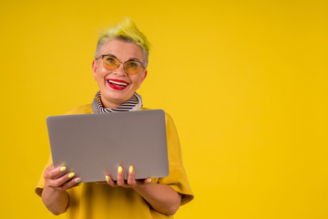 mature old woman with colored stylish yellow dyed hair and red lipstick lips in warm dress holding modern laptop background studio