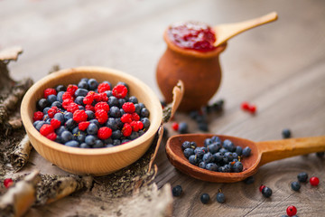 Blueberry and raspberry berry jam in a clay pot, a wooden spoon with fresh berries and birch bark on a wooden background
