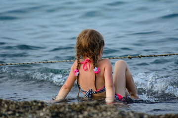 A girl with pigtails sits on the seashore. A wave rushes over her legs. The concept is a happy childhood. Sea holiday