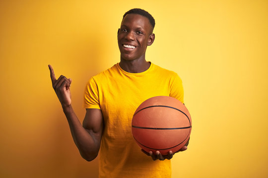 African american athlete man holding basketball ball standing over isolated yellow background very happy pointing with hand and finger to the side