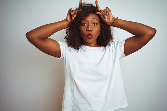 Young african american woman wearing t-shirt standing over isolated white background doing funny gesture with finger over head as bull horns