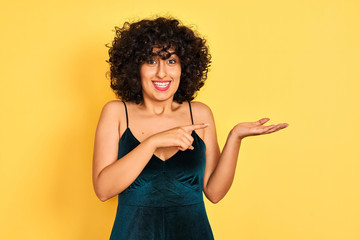 Young arab woman with curly hair wearing elegant dress over isolated yellow background amazed and smiling to the camera while presenting with hand and pointing with finger.