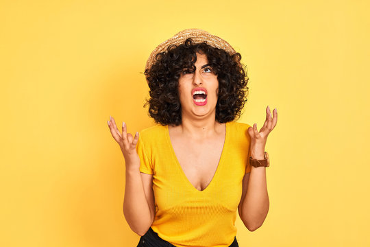 Young arab woman with curly hair wearing t-shirt and hat over isolated yellow background crazy and mad shouting and yelling with aggressive expression and arms raised. Frustration concept.