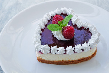 Heart shaped no-bake cheesecake with graham cracker crust, cream cheese filling and raspberry jelly, decorated with whipped cream on white plate.