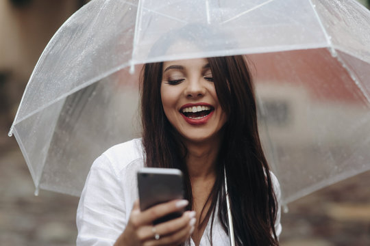 Close up portrait of smiling beautiful girl with umbrella, using smartphone, red wall on the background. Small depth of field. It's summer rain.