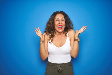 Fototapeta premium Middle age senior woman with curly hair standing over blue isolated background celebrating crazy and amazed for success with arms raised and open eyes screaming excited. Winner concept