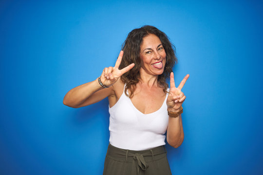 Middle age senior woman with curly hair standing over blue isolated background smiling with tongue out showing fingers of both hands doing victory sign. Number two.