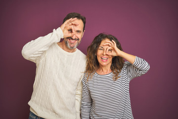 Naklejka premium Beautiful middle age couple wearing winter sweater over isolated purple background doing ok gesture with hand smiling, eye looking through fingers with happy face.