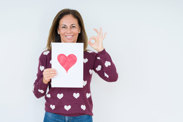 Fototapeta premium Middle age woman holding card gift with red heart over isolated background doing ok sign with fingers, excellent symbol