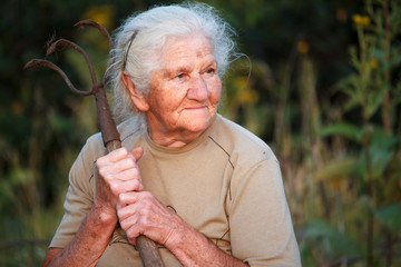 Close-up portrait of an old woman with gray hair holding a rusty pitchfork or chopper in her hands,...
