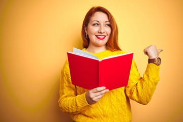 Young redhead teacher woman reading red book over yellow isolated background pointing and showing with thumb up to the side with happy face smiling