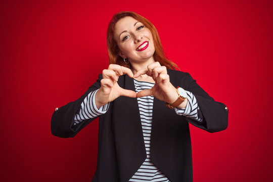 Beautiful redhead business woman wearing elegant jacket over isolated red background smiling in love showing heart symbol and shape with hands. Romantic concept.