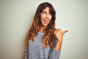 Young beautiful woman wearing stripes t-shirt over white isolated background smiling with happy face looking and pointing to the side with thumb up.