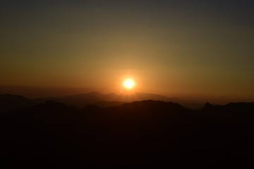 Picture of a sun setting behind a dense forest area followed by mountains.	