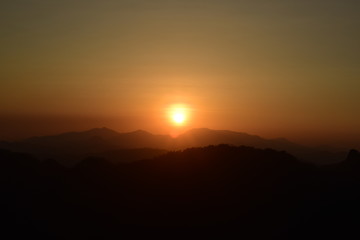 Fototapeta premium sun setting behind a dense forest area followed by mountains.
