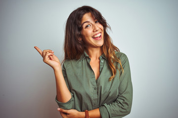 Young beautiful woman wearing green shirt standing over grey isolated background with a big smile on face, pointing with hand finger to the side looking at the camera.