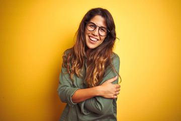 Young beautiful woman wearing green shirt and glasses over yelllow isolated background happy face smiling with crossed arms looking at the camera. Positive person.