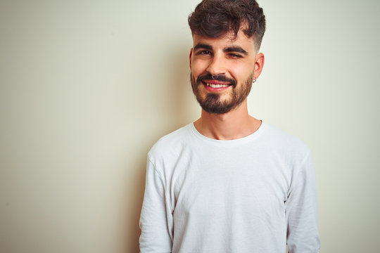 Young man with tattoo wearing t-shirt standing over isolated white background winking looking at the camera with sexy expression, cheerful and happy face.