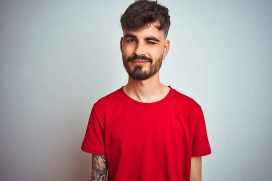 Young man with tattoo wearing red t-shirt standing over isolated white background winking looking at the camera with sexy expression, cheerful and happy face.