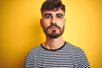 Young man with tattoo wearing striped t-shirt standing over isolated yellow background with serious expression on face. Simple and natural looking at the camera.