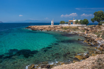 Fototapeta na wymiar The wild coast of Aegina island with clear and blue waters of Mediterranean sea and the old small lighthouse in the background, in Saronic gulf, Greece.