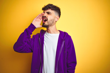 Young man with tattoo wearing sport purple sweatshirt over isolated yellow background shouting and screaming loud to side with hand on mouth. Communication concept.