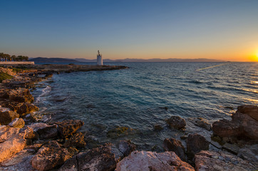 Fototapeta premium Wild coast of the Aegina island and the old small lighthouse in the background, Saronic gulf, Greece, at sunset.