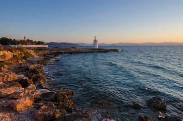 Fototapeta na wymiar Wild coast of the Aegina island and the old small lighthouse in the background, Saronic gulf, Greece, at sunset.