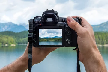 Fototapete Dunkelbraun Hände eines männlichen Fotografen, der eine Digitalkamera hält und eine idyllische Landschaft mit einem See und Bergen fotografiert, während das Bild auf dem Display angezeigt wird