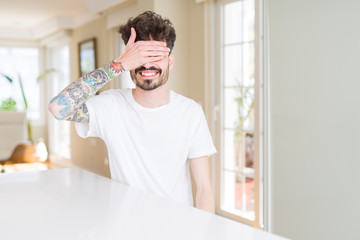 Young man wearing casual t-shirt sitting on white table smiling and laughing with hand on face covering eyes for surprise. Blind concept.