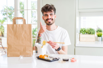Young man eating asian sushi from home delivery happy with big smile doing ok sign, thumb up with fingers, excellent sign