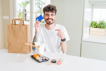 Young man eating asian sushi from home delivery using credit card as payment with surprise face pointing finger to himself