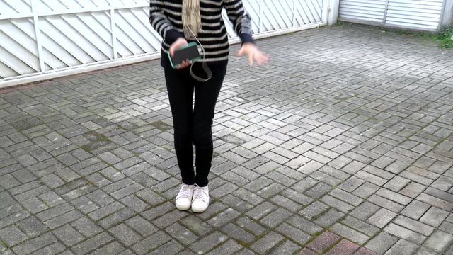 Funny girl dancing and listening music on cellphone on white gate background. Unrecognizable model body holding smartphone and earphones wearing casual black pants and pullover and white sneakers.