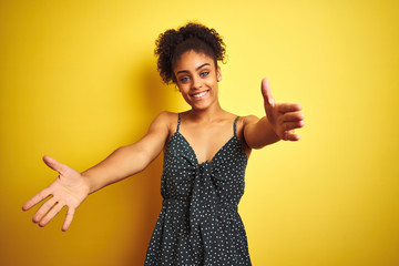 African american woman wearing summer casual green dress over isolated yellow background looking at the camera smiling with open arms for hug. Cheerful expression embracing happiness.