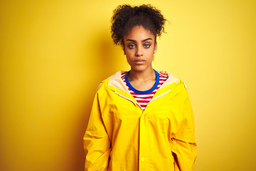 Obraz na płótnie Canvas Young african american woman wearing rain coat over isolated yellow background with serious expression on face. Simple and natural looking at the camera.