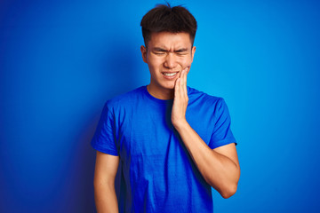 Young asian chinese man wearing t-shirt standing over isolated blue background touching mouth with hand with painful expression because of toothache or dental illness on teeth. Dentist concept.