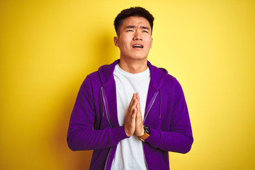 Young asian chinese man wearing purple sweatshirt standing over isolated yellow background begging...