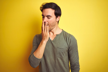 Young handsome man wearing casual t-shirt standing over isolated yellow background bored yawning tired covering mouth with hand. Restless and sleepiness.