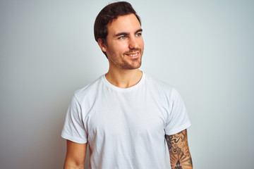 Young handsome man with tattoo wearing casual t-shirt over isolated white background looking away to side with smile on face, natural expression. Laughing confident.