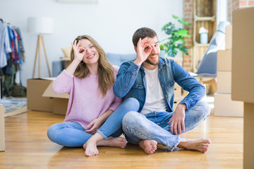 Fototapeta na wymiar Young beautiful couple moving to a new house sitting on the floor doing ok gesture with hand smiling, eye looking through fingers with happy face.