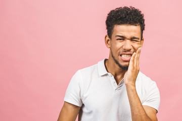 Indoor portrait of young worried African American male isolated on white background dressed in casual experiencing strong toothache pressing hand to chin because of sore jaws.