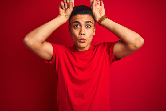 Young brazilian man wearing t-shirt standing over isolated red background doing funny gesture with finger over head as bull horns