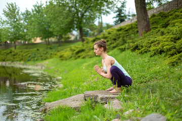 Young beautiful caucasian brunette girl doing yoga on a green lawn against the background of the river